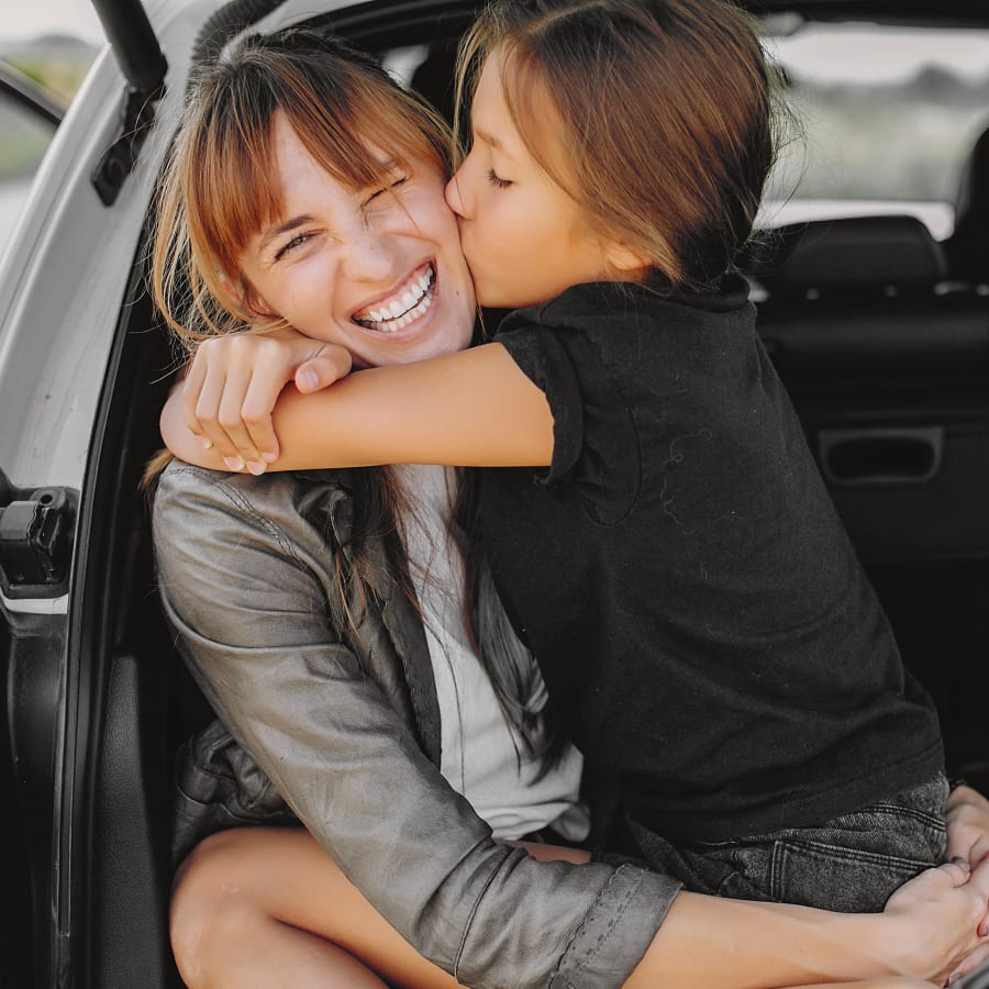 A woman sits in the back of an SUV smilng while a little girl sits on her lap hugging her and giving her a kiss on the cheek.