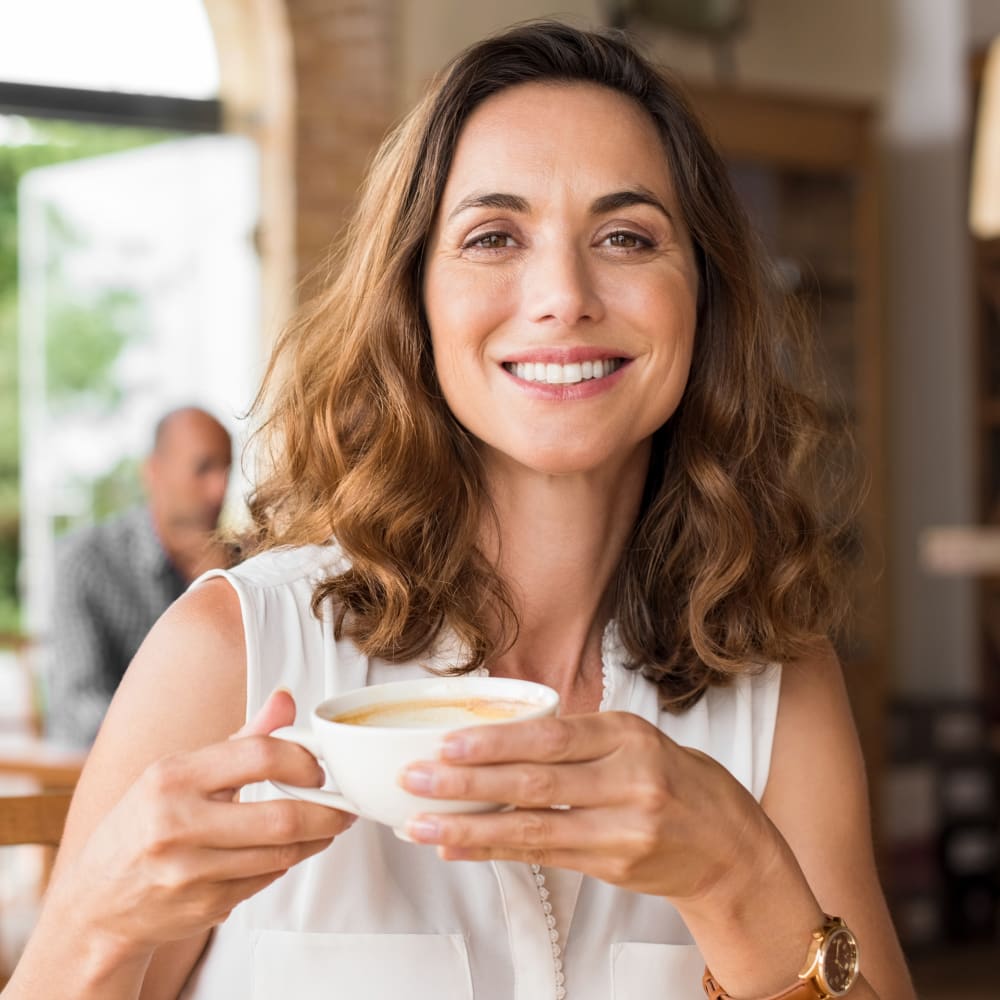 A woman sits at a coffee shop smilng and holding up a cup of coffee.