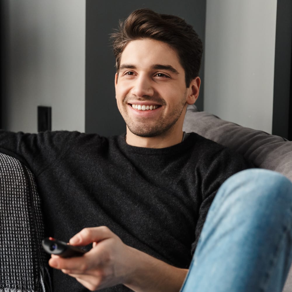 A man in a black t-shirt sits on a sofa holding a remote and smiling while lookign beyond the camera.
