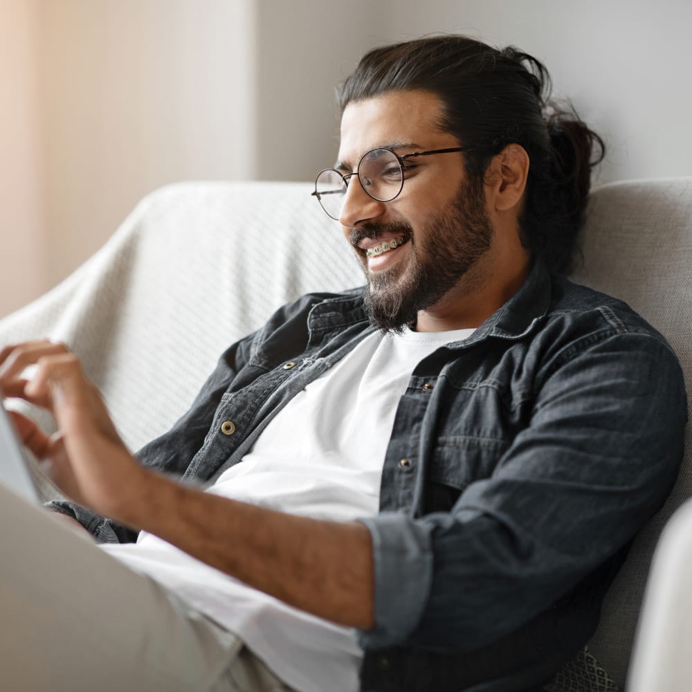 A man with glasses and braces sits on a sofa smiling while using a tablet.