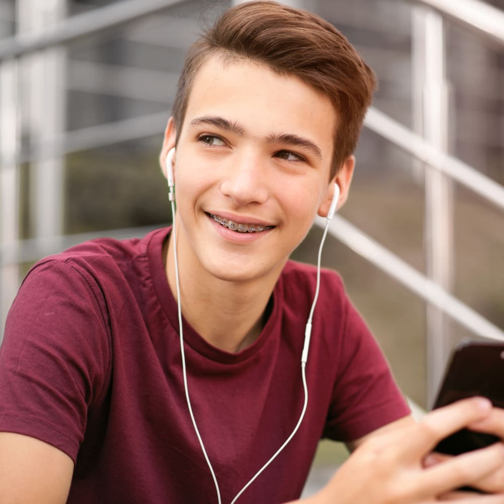 A boy with braces sits outside smiling and looking off to the side while wearing heaphones.