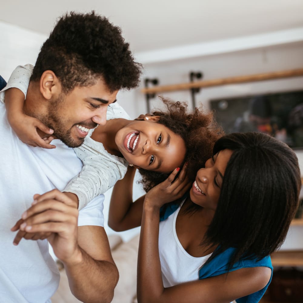 A family stands together while the man has the child up over his shoulder. The little girl is smiling and laughing while the woman holds the girl's head.