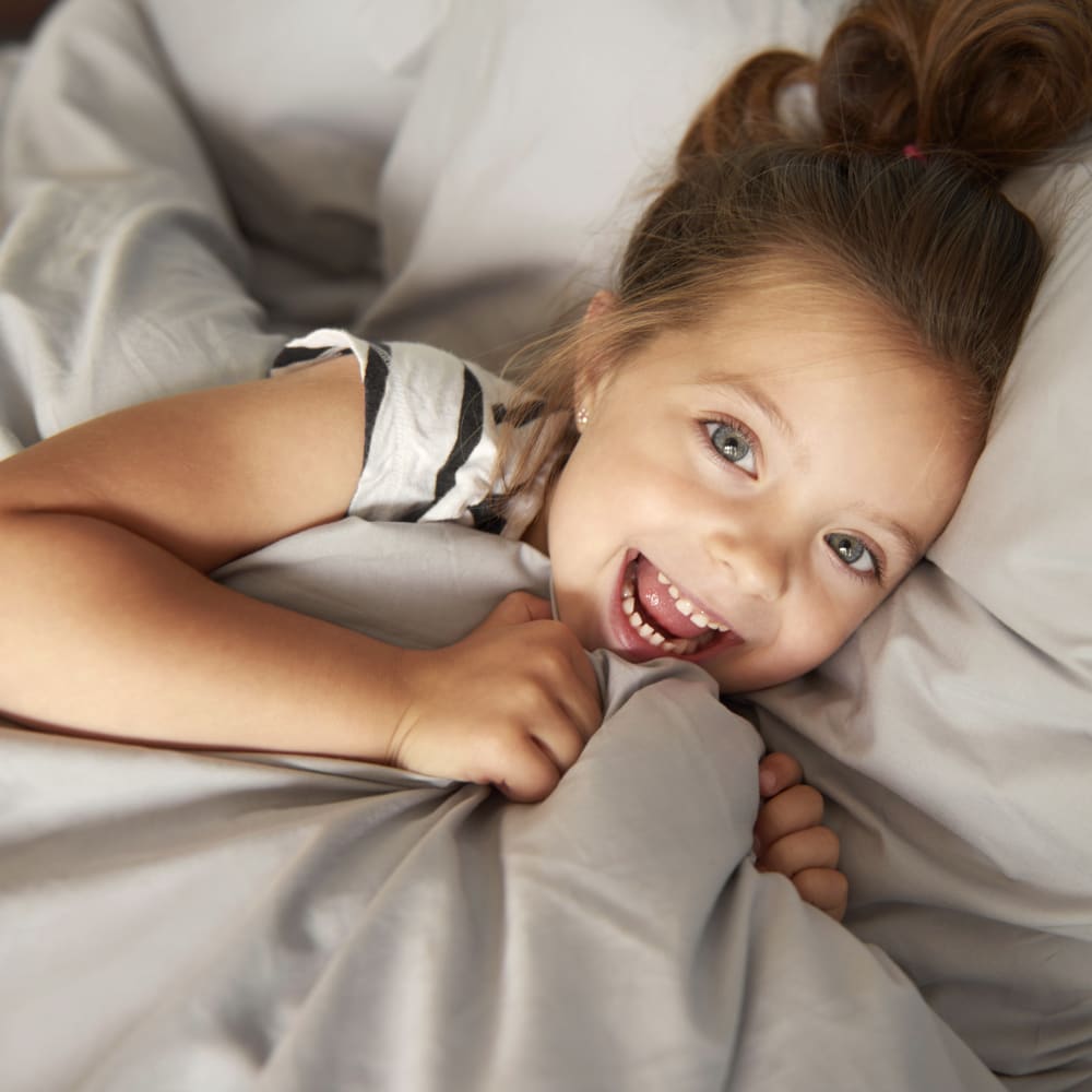A little girl lays in a bed smiling with the blankets pulled up to her chin.