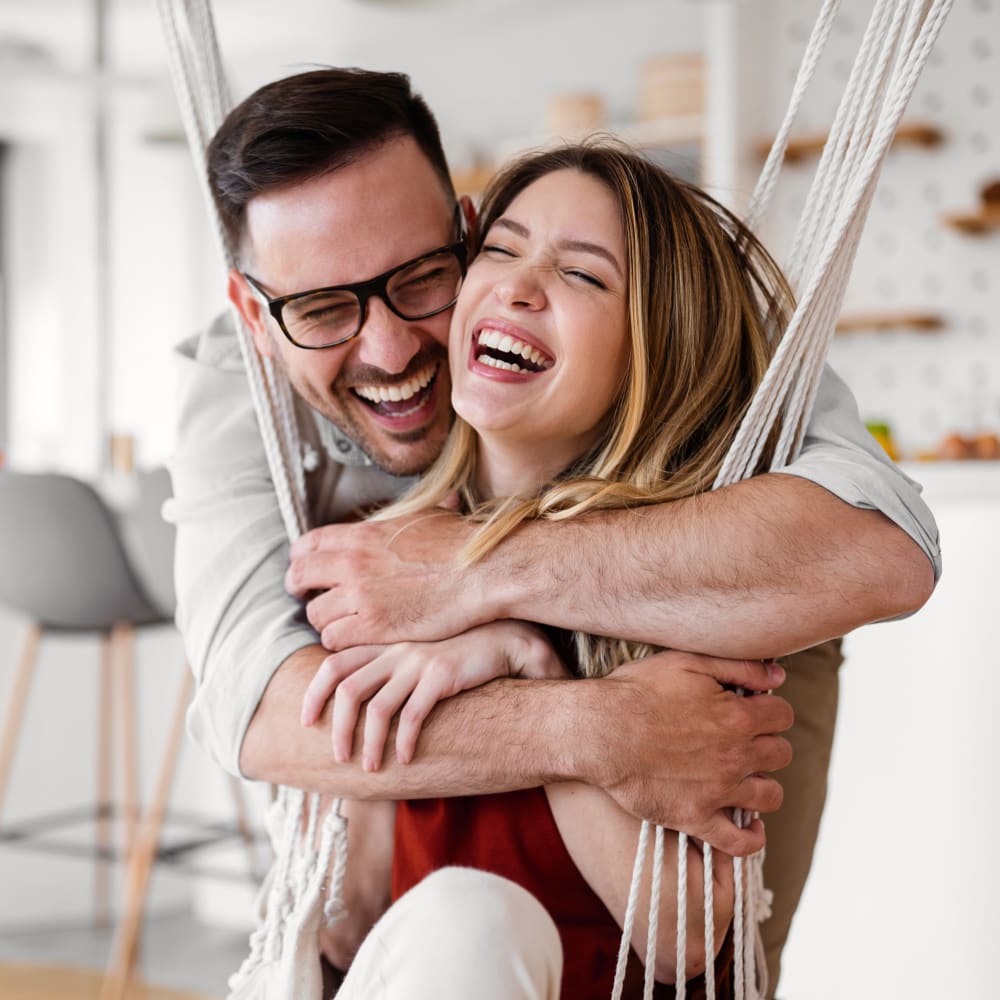 A woman sits on an indoor swing smiling and laughing while a man hugs her from behind.