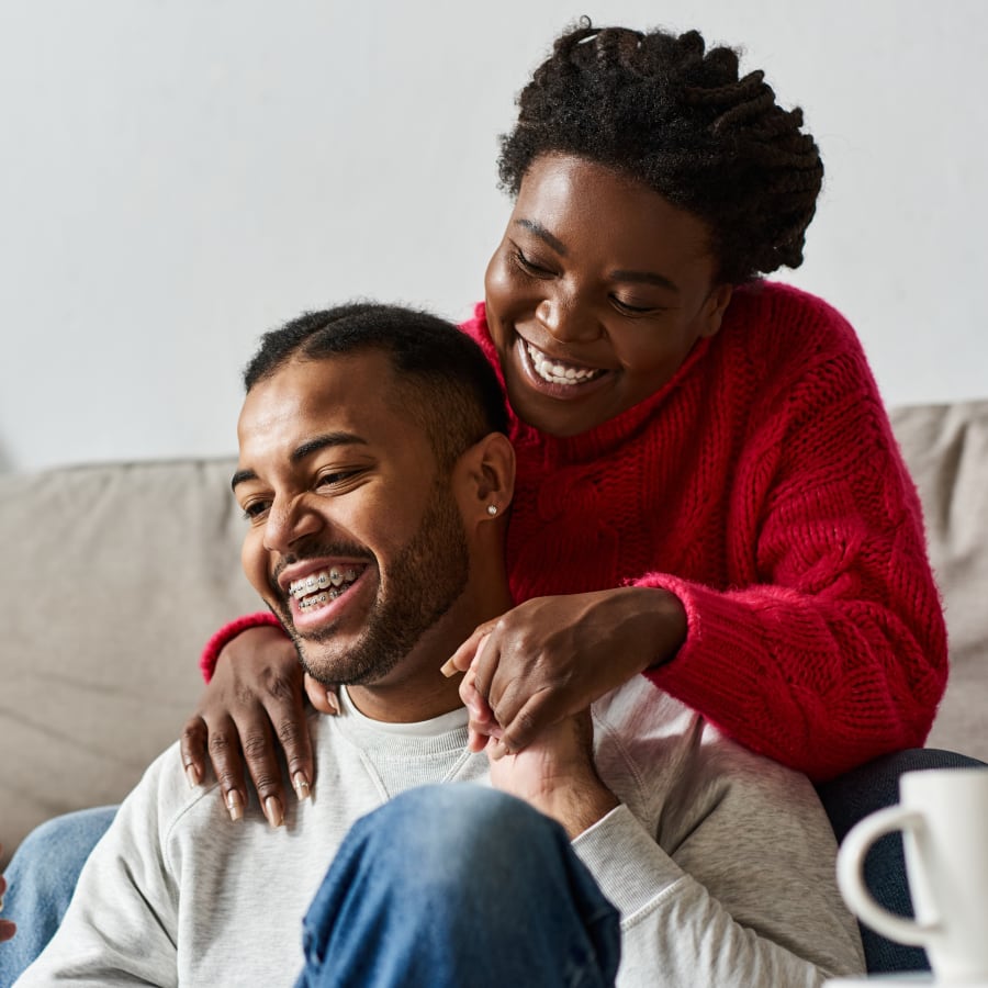 A man with braces sits on the floor looking ahead and smiling while a woman sits on teh sofa behind him and holds his hand and shoulder.