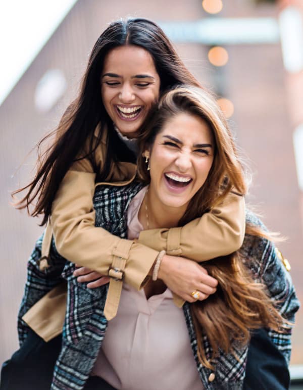 A woman stands outside while carrying a young woman on her back. They are both laughing and smiling.